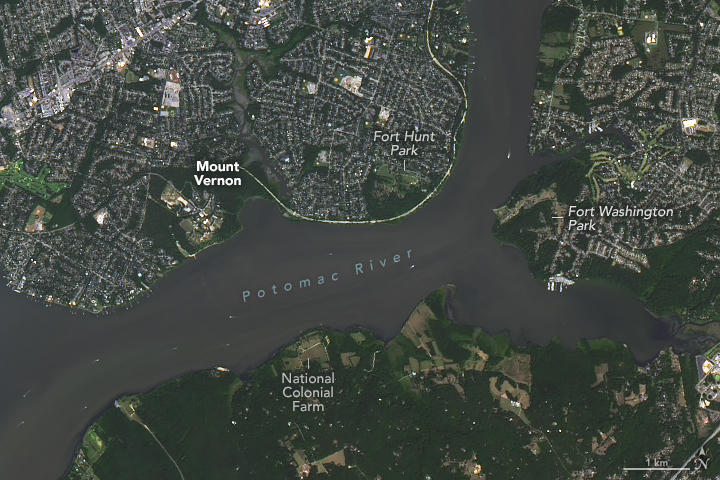 A satellite image shows the Potomac River with Mount Vernon and Fort Hunt Park on the north side of the river and Fort Washington Park and National Colonial Farm on the south side.  Most of the land in this area is gray and covered with development, but Mount Vernon and the parks are green.