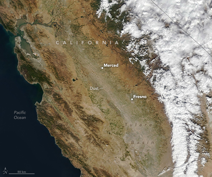 A satellite image of California shows narrow plumes of dust blowing through the Central Valley toward the southeast. The landscape is generally brown but parts of the Central Valley are green with crops. The right edge of the image has many white clouds.